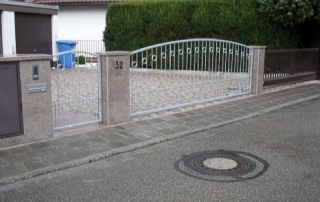 Toranlage bestehend aus einer verzinkten RHP-Rahmen 40x40mm mit senkrecht eingeschweißten Rundstäben d=12mm, inkl. Zierornamenten, inkl. verstellbarer Edestahlscharniere, inkl. elektrischer Toröffnung.